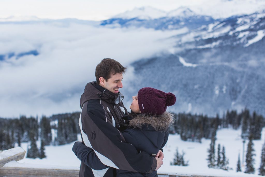 a man and a woman embrace at an engagement session on top of a mountain in whistler